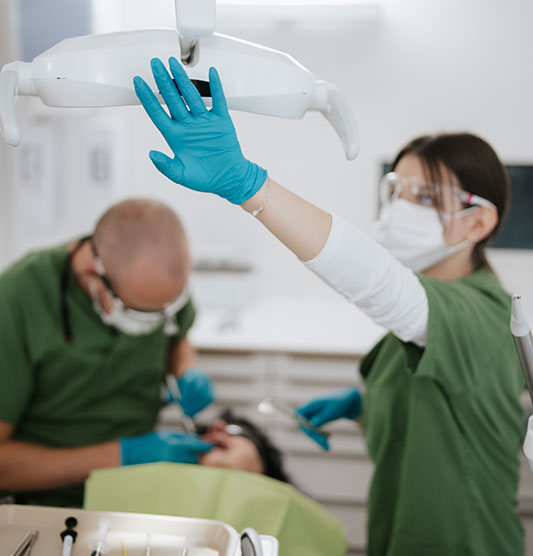 Dr Neubert führt eine Zahnbehandlung an einer Patientin durch. Die Arzthelferin hält die linke Hand nach oben zur Lampe - sie trägt einen türkisen Handschuh