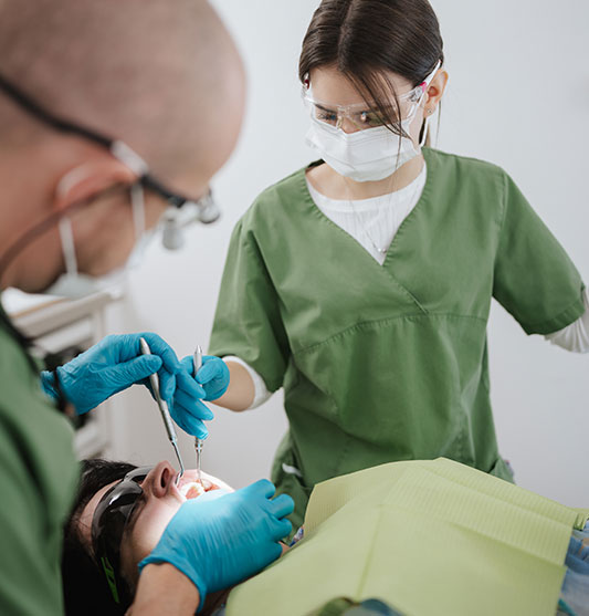 Dr Neubert führt eine Zahnbehandlung an einer Patientin durch. Ihm gegenüber steht die Arzthelferin und assistiert ihr