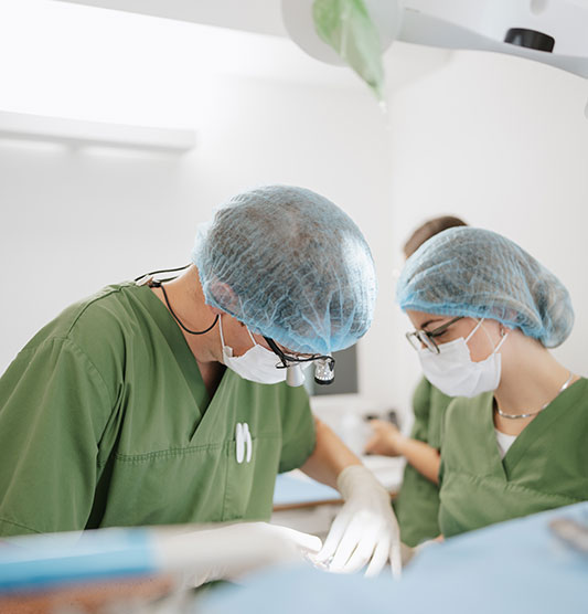 Dr Schipp führt eine Zahnoperation durch. Gegenüber von ihm steht eine Arzthelferin, die ihm assistiert. Beide tragen Mundschutz und eine blaue Haube